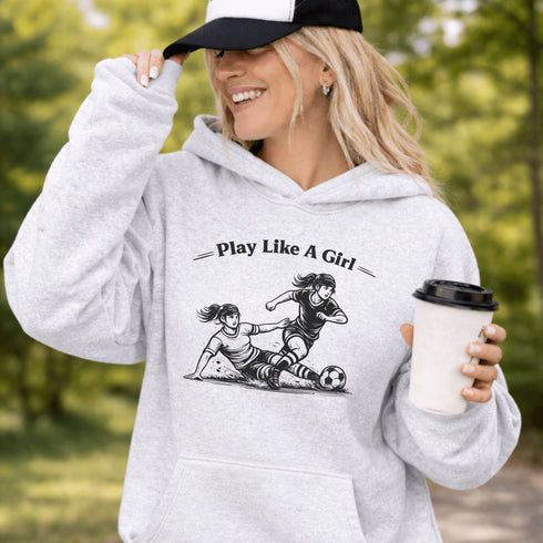 Woman wearing a grey Play Like A Girl soccer hoodie with girls soccer graphic, standing on a soccer field with hands in pocket