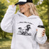 Woman wearing a grey Play Like A Girl soccer hoodie with girls soccer graphic, standing on a soccer field with hands in pocket