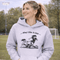 Woman wearing a grey Play Like A Girl soccer hoodie with girls soccer graphic, standing on a soccer field with hands in pocket