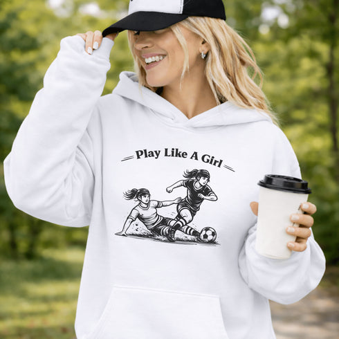 Woman wearing a white Play Like A Girl soccer hoodie with girls soccer graphic, standing on a soccer field with hands in pocket