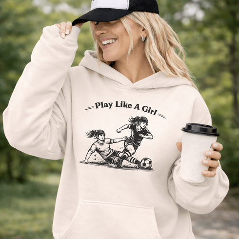 Woman wearing a light tan Play Like A Girl soccer hoodie with girls soccer graphic, standing on a soccer field with hands in pocket