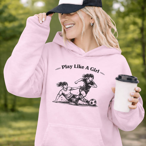 Woman wearing a light pink Play Like A Girl soccer hoodie with girls soccer graphic, standing on a soccer field with hands in pocket