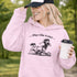 Woman wearing a light pink Play Like A Girl soccer hoodie with girls soccer graphic, standing on a soccer field with hands in pocket