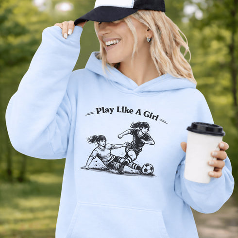 Woman wearing a light blue Play Like A Girl soccer hoodie with girls soccer graphic, standing on a soccer field with hands in pocket