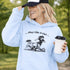 Woman wearing a light blue Play Like A Girl soccer hoodie with girls soccer graphic, standing on a soccer field with hands in pocket