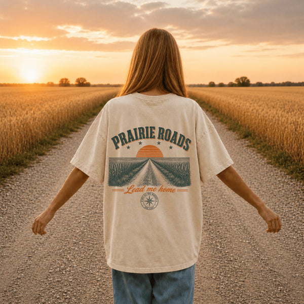 Back view of a person wearing a cream Comfort Colors t-shirt featuring a vintage prairie road and wheat field sunset graphic while standing on a gravel road between golden wheat fields at sunset.