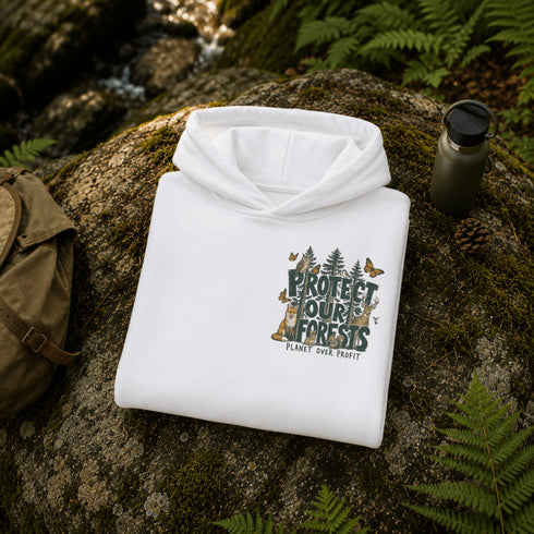 A folded white hoodie with a forest-themed graphic reading 'PROTECT OUR FORESTS' and 'PLANET OVER PROFIT' is placed on a mossy rock outdoors, surrounded by ferns, with a backpack and water bottle nearby.