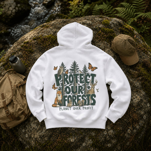 A white pullover hoodie laid flat on a mossy rock outdoors, with a large forest-themed back graphic reading “PROTECT OUR FORESTS” and “PLANET OVER PROFIT,” plus illustrated animals and butterflies. A tan cap, backpack, and water bottle are placed around it in a natural outdoor setting.