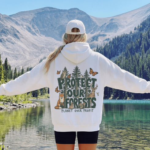 A woman stands with her back to the camera in front of a mountain lake, wearing a white hoodie with a large back graphic that reads 'PROTECT OUR FORESTS PLANET OVER PROFIT' with trees, butterflies, and animals, and a white cap.