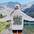 A woman stands with her back to the camera in front of a mountain lake, wearing a white hoodie with a large back graphic that reads 'PROTECT OUR FORESTS PLANET OVER PROFIT' with trees, butterflies, and animals, and a white cap.