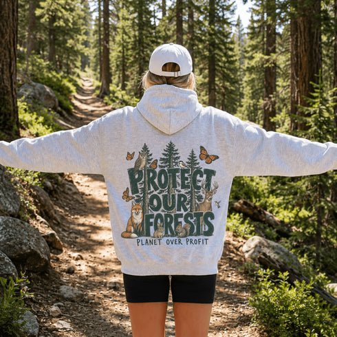 A person standing on a forest trail wearing a Light Grey hoodie with a large back graphic that reads “PROTECT OUR FORESTS” and “PLANET OVER. PROFIT,” along with trees, a fox, butterflies, and deer.