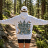 A person standing on a forest trail wearing a Light Grey hoodie with a large back graphic that reads “PROTECT OUR FORESTS” and “PLANET OVER. PROFIT,” along with trees, a fox, butterflies, and deer.