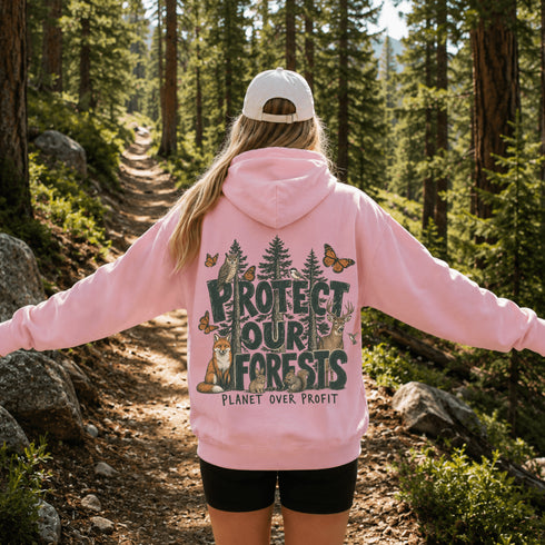 A woman standing on a forest trail viewed from behind, wearing a pink hoodie with a large back graphic that reads “PROTECT OUR FORESTS” and “PLANET OVER PROFIT,” along with trees, butterflies, a fox, and a white cap.