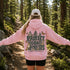 A woman standing on a forest trail viewed from behind, wearing a pink hoodie with a large back graphic that reads “PROTECT OUR FORESTS” and “PLANET OVER PROFIT,” along with trees, butterflies, a fox, and a white cap.