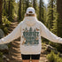 A person standing on a forest trail viewed from behind, wearing a cream hoodie with a large back graphic that reads “PROTECT OUR FORESTS” and “PLANET OVER PROFIT,” along with trees, butterflies, a fox, and deer. The person also wears a white cap and black shorts.