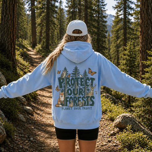 A person standing on a forest trail, viewed from behind, wearing a light blue hoodie with a large back graphic that reads “PROTECT OUR FORESTS” and “PLANET OVER PROFIT,” along with forest animals and butterflies. The person is also wearing a white cap and black shorts.