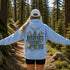 A person standing on a forest trail, viewed from behind, wearing a light blue hoodie with a large back graphic that reads “PROTECT OUR FORESTS” and “PLANET OVER PROFIT,” along with forest animals and butterflies. The person is also wearing a white cap and black shorts.