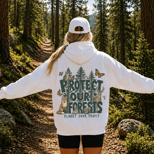 A woman stands on a forest trail with her back to the camera, wearing a white hoodie with a large back graphic that reads “PROTECT OUR FORESTS” and “PLANET OVER PROFIT,” along with woodland animals, trees, and butterflies. She is also wearing a white cap and black shorts.