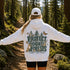 A woman stands on a forest trail with her back to the camera, wearing a white hoodie with a large back graphic that reads “PROTECT OUR FORESTS” and “PLANET OVER PROFIT,” along with woodland animals, trees, and butterflies. She is also wearing a white cap and black shorts.