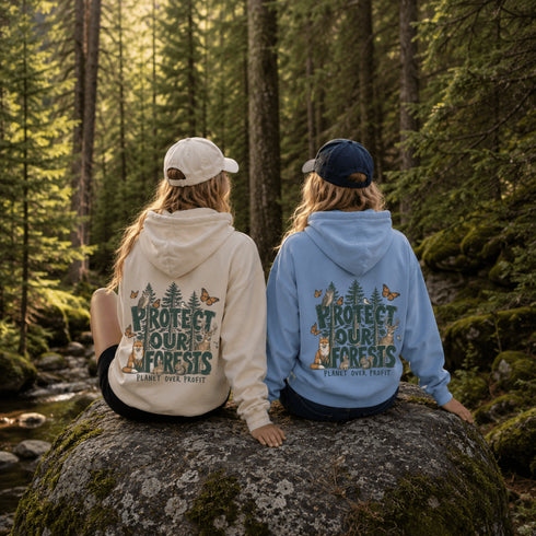 Two people sitting on a mossy rock in a forest, seen from behind, wearing matching hoodies with a back graphic that reads “PROTECT OUR FORESTS PLANET OVER PROFIT.” One hoodie is cream and the other is light blue, with the models wearing a white cap and a navy cap respectively.