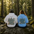 Two people sitting on a mossy rock in a forest, seen from behind, wearing matching hoodies with a large back graphic that reads “PROTECT OUR FORESTS” and “PLANET OVER PROFIT,” one in white and one in light blue.