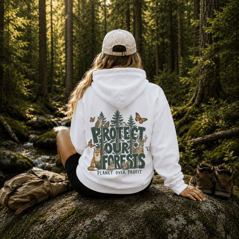 A woman sits on a mossy rock in a forest, viewed from behind, wearing a white hoodie with a large back graphic that reads “PROTECT OUR FORESTS” and “PLANETY OVER PROFIT,” surrounded by trees, butterflies, and animals. A tan backpack rests on the rock beside her, with hiking boots nearby.