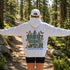 A person standing on a forest trail wearing a light heather gray hoodie from the back, which features a large graphic print reading 'PROTECT OUR WILDLIFE' and 'LEARN BEFORE YOU BURN' with animals and butterflies. The person has their arms outstretched and is wearing a white cap and black shorts.