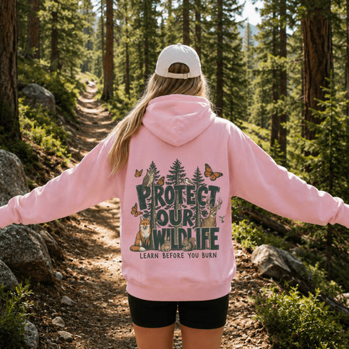 A woman viewed from behind on a forest trail wearing a light pink hoodie with a large back graphic that reads “PROTECT OUR WILDLIFE” and “LEARN BEFORE YOU BURN,” along with trees, animals, and butterflies. She is also wearing a white cap and black shorts.