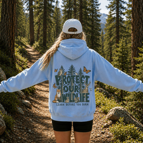 A woman standing on a forest trail seen from behind, wearing a light blue hoodie with a large back graphic that reads “PROTECT OUR WILDLIFE” and “LEARN BEFORE YOU BURN,” along with deer, trees, butterflies, and a fox. She is also wearing a white cap and black shorts.