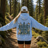 A woman standing on a forest trail seen from behind, wearing a light blue hoodie with a large back graphic that reads “PROTECT OUR WILDLIFE” and “LEARN BEFORE YOU BURN,” along with deer, trees, butterflies, and a fox. She is also wearing a white cap and black shorts.