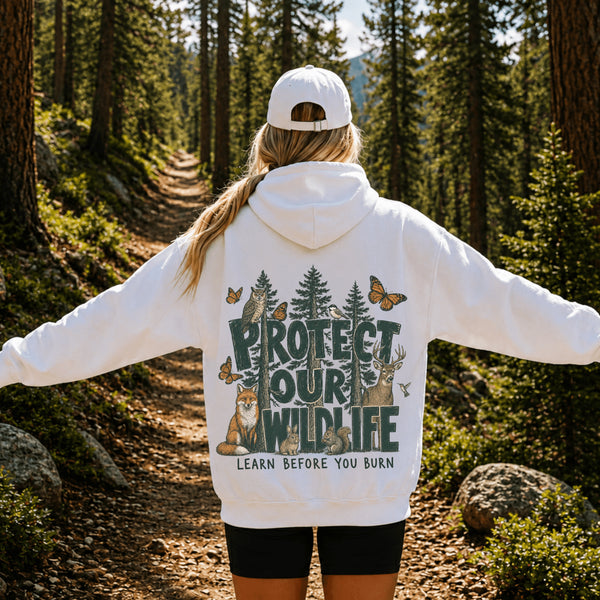 A woman standing on a forest trail viewed from behind, wearing a white hoodie with a large back graphic that reads “PROTECT OUR WILDLIFE” and “LEARN BEFORE YOU BURN,” along with trees, animals, and butterflies. She is also wearing a white cap and black shorts.