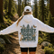 A woman standing on a forest trail viewed from behind, wearing a white hoodie with a large back graphic that reads “PROTECT OUR WILDLIFE” and “LEARN BEFORE YOU BURN,” along with trees, animals, and butterflies. She is also wearing a white cap and black shorts.