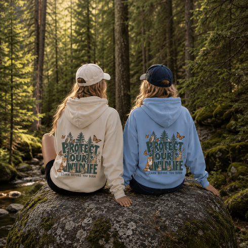 Two people sitting on a large mossy rock in a forest, seen from behind, wearing matching hoodies with a nature-themed 'PROTECT OUR WILDLIFE' graphic on the back. One hoodie is cream and the other is light blue, both with different colored caps.