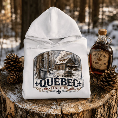 White hoodie with Québec maple syrup cabin design on a wooden stump with maple syrup bottle and pinecones.