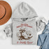 This hoodie features a raccoon—kicked back against a tree stump, wearing a cowboy hat, and looking like it just clocked out of life’s gym membership.