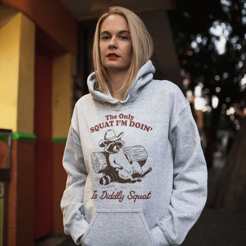 This hoodie features a raccoon—kicked back against a tree stump, wearing a cowboy hat, and looking like it just clocked out of life’s gym membership.