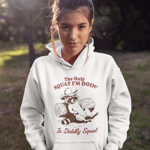 This hoodie features a raccoon—kicked back against a tree stump, wearing a cowboy hat, and looking like it just clocked out of life’s gym membership.