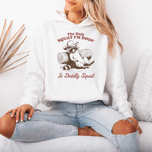 This hoodie features a raccoon—kicked back against a tree stump, wearing a cowboy hat, and looking like it just clocked out of life’s gym membership.