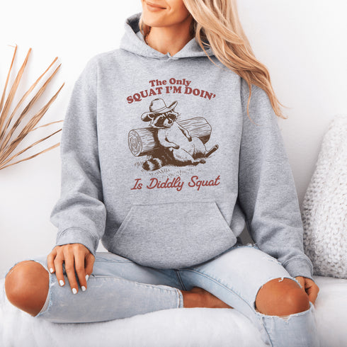 This hoodie features a raccoon—kicked back against a tree stump, wearing a cowboy hat, and looking like it just clocked out of life’s gym membership.