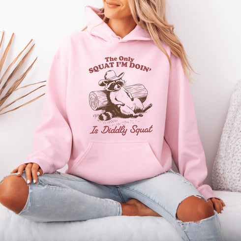 This hoodie features a raccoon—kicked back against a tree stump, wearing a cowboy hat, and looking like it just clocked out of life’s gym membership.