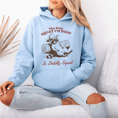 This hoodie features a raccoon—kicked back against a tree stump, wearing a cowboy hat, and looking like it just clocked out of life’s gym membership.