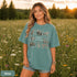 Vintage wildlife graphic on a pepper Comfort Colors t-shirt featuring a bear, fox, owl, squirrel, rabbit, deer, beaver, and lynx with the phrase "Respect the Locals," modeled in a wildflower meadow at sunset.