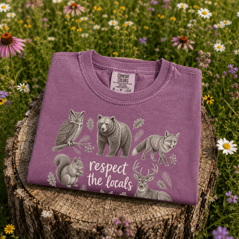 Vintage wildlife graphic on a pepper Comfort Colors t-shirt featuring a bear, fox, owl, squirrel, rabbit, deer, beaver, and lynx with the phrase "Respect the Locals," modeled in a wildflower meadow at sunset.