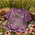 Vintage wildlife graphic on a pepper Comfort Colors t-shirt featuring a bear, fox, owl, squirrel, rabbit, deer, beaver, and lynx with the phrase "Respect the Locals," modeled in a wildflower meadow at sunset.