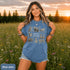 Vintage wildlife graphic on a pepper Comfort Colors t-shirt featuring a bear, fox, owl, squirrel, rabbit, deer, beaver, and lynx with the phrase "Respect the Locals," modeled in a wildflower meadow at sunset.