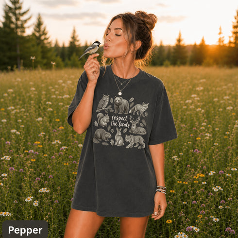 Vintage wildlife graphic on a pepper Comfort Colors t-shirt featuring a bear, fox, owl, squirrel, rabbit, deer, beaver, and lynx with the phrase "Respect the Locals," modeled in a wildflower meadow at sunset.