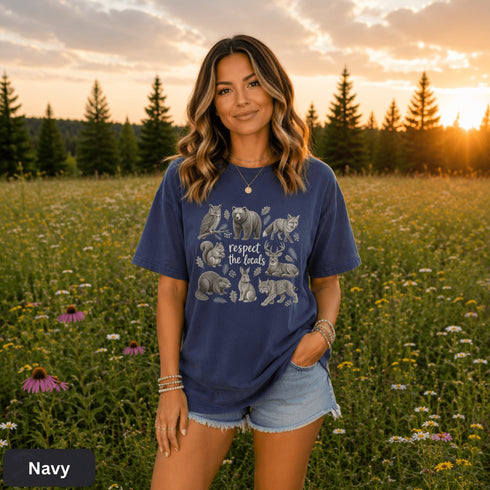 Vintage wildlife graphic on a pepper Comfort Colors t-shirt featuring a bear, fox, owl, squirrel, rabbit, deer, beaver, and lynx with the phrase "Respect the Locals," modeled in a wildflower meadow at sunset.
