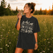 Vintage wildlife graphic on a pepper Comfort Colors t-shirt featuring a bear, fox, owl, squirrel, rabbit, deer, beaver, and lynx with the phrase "Respect the Locals," modeled in a wildflower meadow at sunset.