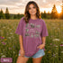 Vintage wildlife graphic on a pepper Comfort Colors t-shirt featuring a bear, fox, owl, squirrel, rabbit, deer, beaver, and lynx with the phrase "Respect the Locals," modeled in a wildflower meadow at sunset.