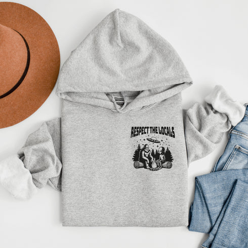 Our Respect the Locals Hoodie features the ultimate forest hangout: Bigfoot and an alien, just two mysterious dudes chilling on logs around a crackling campfire.